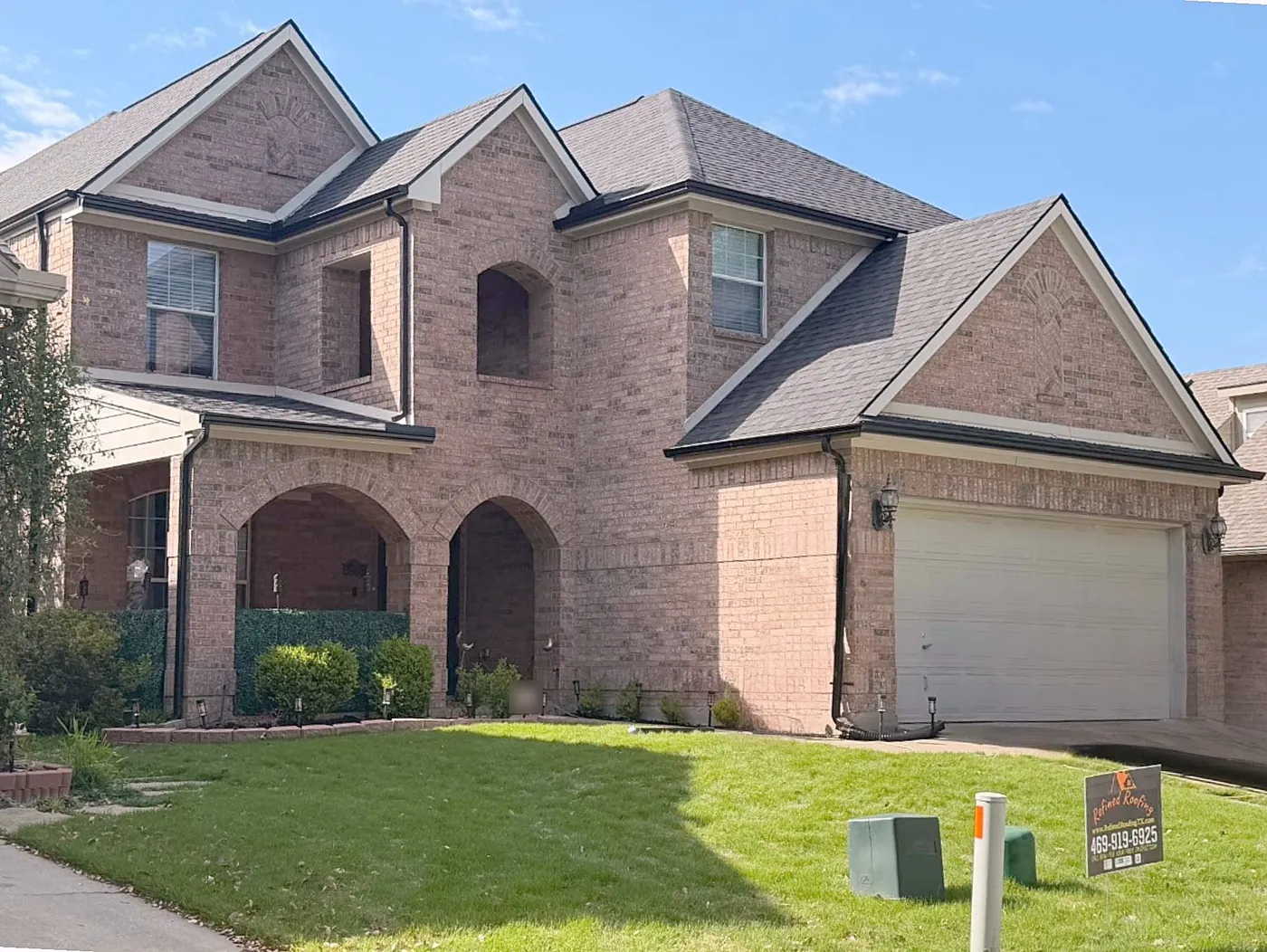 A two-story brick home with a dark shingle roof and a Refined Roofing yard sign in the lawn