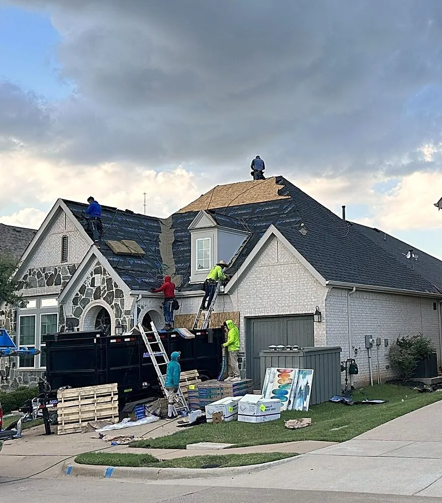 A roofing crew replacing the roof on a stone and white brick home, with a dump trailer and materials in the driveway